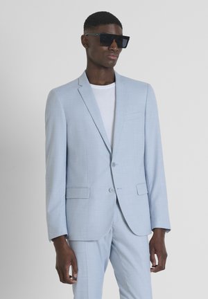 Un costume bleu clair avec un motif à carreaux subtil, doté d'un col cranté, d'une fermeture à un bouton et de poches latérales. Associé à des lunettes de soleil noires.