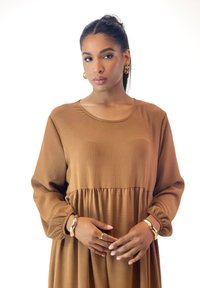Braunes Langarmkleid mit Rundhalsausschnitt und geraffter Taille. Aus glattem Stoff mit dezenter glänzender Optik, accessoriert mit goldenen Ohrringen und Schmuck.