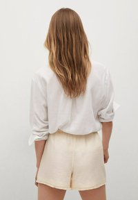 Chemise en lin blanc avec manches retroussées, associée à un short de couleur crème avec un ourlet effiloché. Modèle vu de dos.