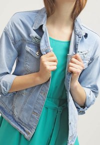 Veste en denim bleu clair avec des détails usés, devant boutonné et poches à la poitrine. Portée par-dessus une robe turquoise avec une taille cintrée.