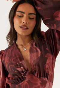 Röd blommig omlottklänning med djup V-ringning, draperad midja och långa ballongärmar. Silverhalsband med blomformad hängsmycke ger extra detalj.