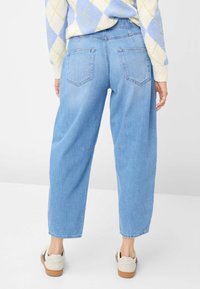 Lichtblauwe, hoogwater-spijkerbroek met een losse pasvorm, voorzien van achterzakken en een onafgewerkte zoom. Gecombineerd met crèmekleurige sneakers op een neutrale achtergrond.