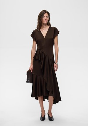 Frau trägt ein dunkelbraunes asymmetrisches Kleid mit Rüschen, schwarze spitze Schuhe, silbernes Armband und hält eine kleine dunkle Handtasche.