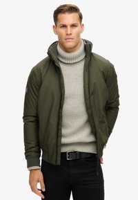 Olivgrüner Bomberjacke mit hohem Kragen, Reißverschluss und gerippten Bündchen, getragen über einem hellgrauen grob gestrickten Pullover. Schwarze Hose vervollständigt den Look.