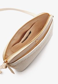 Beżowa torba na ramię z teksturowaną powierzchnią, złotym suwakiem i przestronnym jasnym wnętrzem z małą kieszonką z metką.