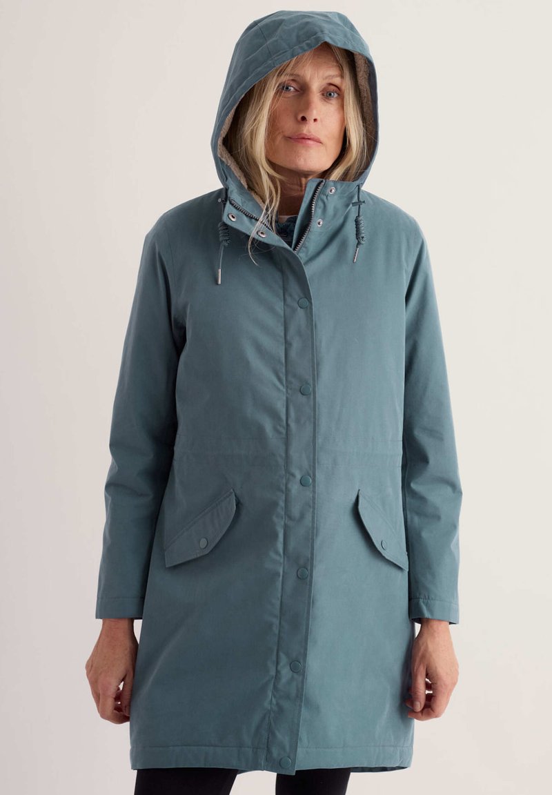 Femme aux longs cheveux blonds porte un imperméable à capuche bleu canard avec des boutons devant, des poches à rabat et des cordons, debout devant un fond clair.