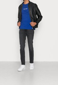 Czarna skórzana kurtka bombowca na niebieskiej koszulce Calvin Klein, zestawiona z dopasowanymi szarymi dżinsami i białymi trampkami, stojąca na neutralnym tle.