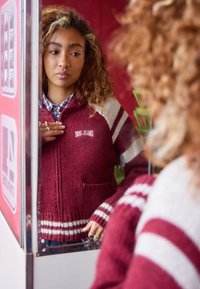 Young woman with curly hair wears a maroon and cream striped sweater, looking thoughtfully at her reflection in a mirror.