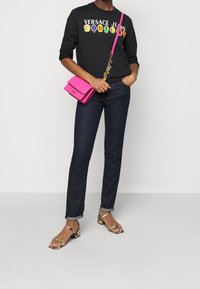 Svart sweatshirt med färgglad "Versace Jeans Couture"-logotyp, mörkblå jeans, rosa crossbody-väska och mönstrade klacksandaler.