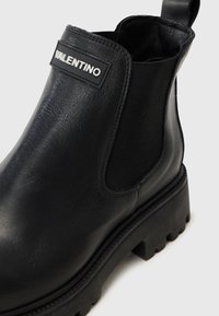 Black leather ankle boot with elastic side panel, chunky tread sole, and a branded patch on the front, showcasing a smooth texture.