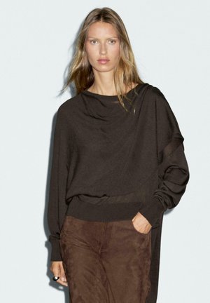 Brown oversized sweater with a draped neckline and long sleeves. Paired with brown suede pants. Smooth texture and relaxed fit.