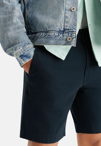 Jachetă din denim cu spălare deschisă, design cu nasturi; suprapusă peste o cămașă verde mentă. Șorturi negre cu o textură fină și tiv curat.
