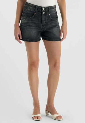 Femme portant un short en jean noir avec des poches avant, une fermeture à boutons, un haut blanc et des sandales blanches à talons, debout devant un fond uni.