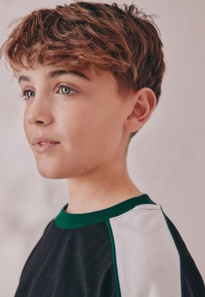 Fiatal személy rövid, hullámos barna hajjal, fekete pólót visel fehér és zöld díszítéssel, enyhén balra néz egy sima háttér előtt.