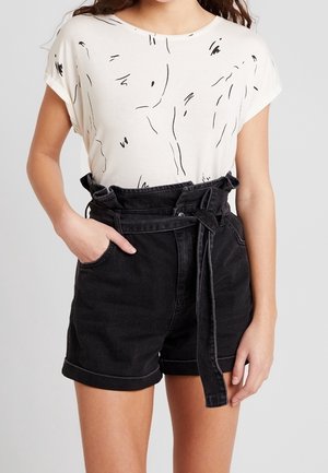 Femme portant un haut blanc à manches courtes avec des lignes noires abstraites et un short en jean noir taille haute avec une ceinture nouée.