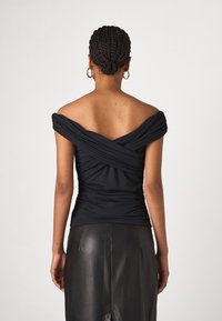 Nainen, jolla on letitetyt hiukset, pukeutuneena olkapäät paljastavaan mustaan yläosaan ja mustaan nahkahameeseen, näkyy selkäpuolelta yksinkertaista taustaa vasten.