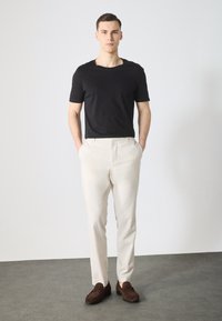 Schwarzfarbenes Crew-Neck T-Shirt, beiges, maßgeschneidertes Hosen, braune Wildleder-Loafer; neutraler Hintergrund betont modernes, einfaches Design.