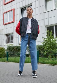 Černá a červená bomber bunda, bílý tričko s grafikou, volné modré džíny a černé tenisky s oranžovými akcenty, zasazené proti pozadí budovy.