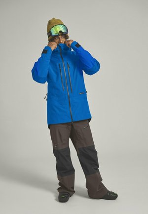 Blue insulated jacket with zippered pockets, paired with brown and black snow pants. Wearing a brown knit hat and mirrored goggles. Black footwear.