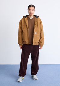 Bruine gewatteerde jas met een zwarte voering, bruin T-shirt en bordeauxrode corduroy broek. Grijze sneakers met groene accenten. Eenvoudig ontwerp, casual pasvorm.