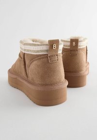 Rudos suede užkulnių batai su pūkinėmis pamušalu, turintys tekstūruotą grietinėlės ir aukso spalvos kraštą bei plokščią gumos padą. Logotipas ant užpakalinės juostos.