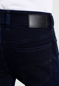 Jeans de mezclilla de color azul oscuro con una textura suave, diseño clásico de cinco bolsillos y un parche rectangular de cuero negro con el logo "DIESEL" en relieve.