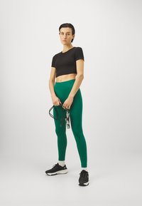 Juoda trumpa marškinėliai, žalia aukšta talija kelnaitės ir juodi sportiniai bateliai su baltais akcentais. Laikydama šokinėjimo virvę, stovi neutralioje fone.