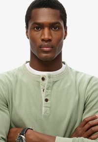 Maglia henley a maniche lunghe di colore verde chiaro con un motivo testurizzato, dotata di scollo a girocollo, tre bottoni marroni e una maglietta interna bianca.