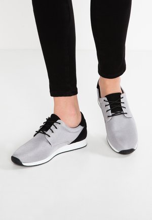 Piedi che indossano scarpe da ginnastica basse grigie e nere con lacci neri, abbinate a pantaloni aderenti neri su un pavimento bianco.