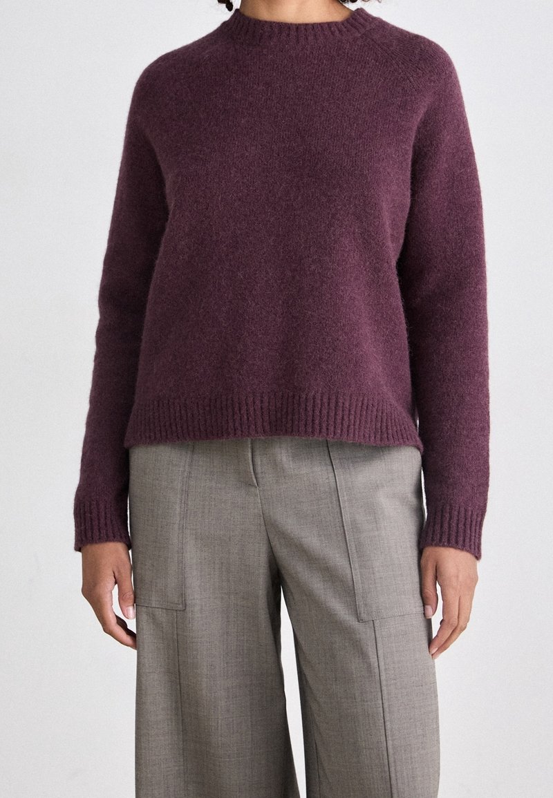 Personne portant un pull en laine violet à manches longues et col rond, ainsi qu'un pantalon gris clair ample taille haute avec des poches avant visibles.