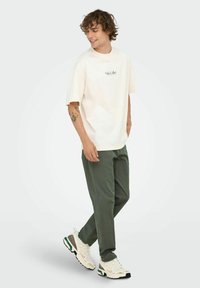 Joven con camiseta oversized color crema, pantalones verde oliva y zapatillas blancas, de pie con una pierna cruzada sobre la otra y sonriendo.