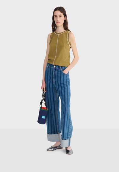 Žena v bezrukávovom olivovozelenom topu s vertikálnym švom, širokých modrých rifliach s prúžkami a manžetami, v kovových balerínkach, držiaca malú námornícky modrú kabelku s červeným vnútrom.