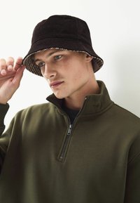 Black corduroy bucket hat with a checkered pattern inside, paired with an olive green zip-up fleece top with a high collar and smooth texture.