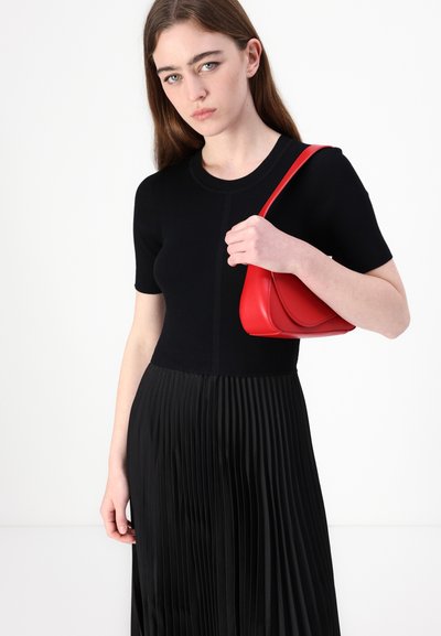 Top en maille noire à côtes avec des manches courtes, associé à une jupe plissée noire. Le modèle tient un sac à main en cuir rouge lisse.