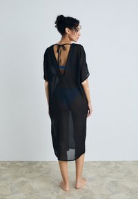Black sheer cover-up dress with short sleeves, V-back design, and tie closure. Length reaches mid-calf, revealing a blue swimsuit underneath.