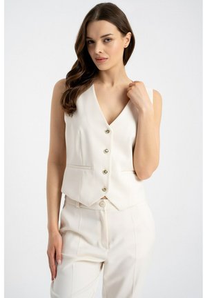 Kobieta o długich ciemnych włosach, ubrana w białą, bezrękawnikową kamizelkę z guzikami i dopasowane spodnie z wysokim stanem, na jednolitym tle.