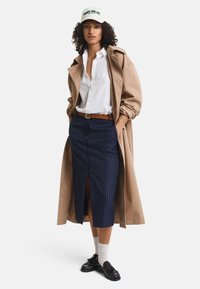 Beiger Trenchcoat, weißes Hemd mit Knöpfen, navyblauer Rock mit Nadelstreifen, brauer Gürtel und dunkle Schuhe mit Socken. Weiße Mütze mit grüner Schrift.