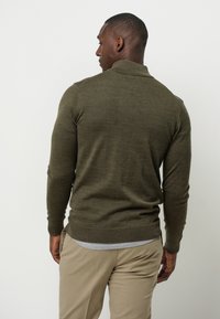 Olivengrüner Pullover mit gerippten Bündchen und Saum, schickem Kragen, taillierter Passform und leichtem Strickmaterial, getragen mit einem hellgrauen Unterhemd und beigen Hosen.