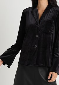 Czarna aksamitna koszula zapinana na guziki z kieszenią na piersi, kołnierzykiem z wycięciem i długimi rękawami, o fakturowanym wykończeniu i rozszerzanych mankietach.