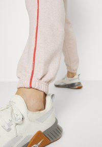 Gros plan d'une personne portant un jogger rose clair avec des bandes latérales rouges et des chaussures de sport blanches avec des semelles grises et orange.