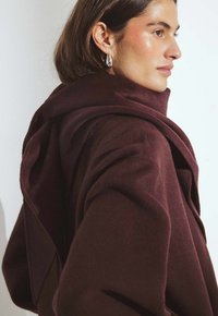 Il cappotto in lana color bordeaux presenta un colletto drappeggiato, una texture morbida e una silhouette rilassata. Il tessuto ha una finitura liscia e dettagli minimi.