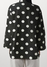 Chemise noire à pois blancs, dotée d'un col, de manches longues et d'une coupe décontractée. Le tissu semble lisse avec des détails de couture visibles.