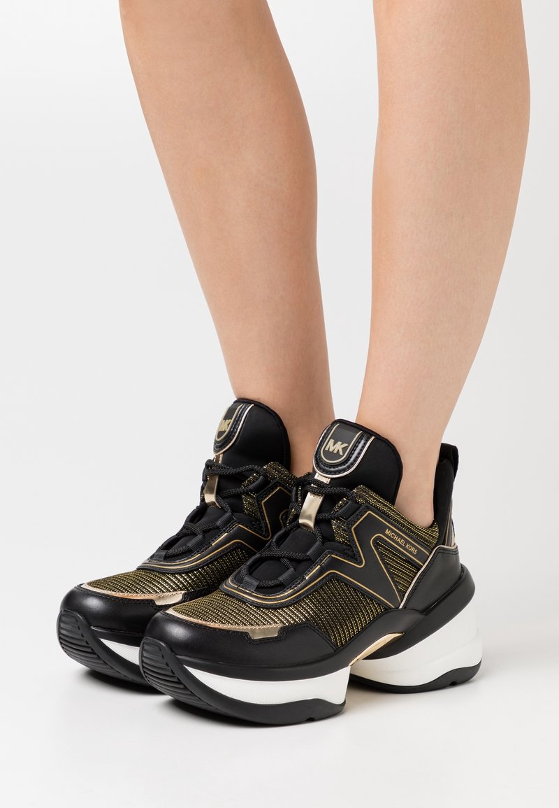 Schwarze und goldene Sportschuhe mit einem strukturierten Mesh-Obermaterial, dicker weißer Sohle und goldenen Akzenten. Schnürdesign mit hohem Kragen.