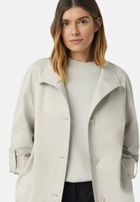 Trench-coat gris clair avec un large col, fermeture à boutons, et des manches texturées. Porté par-dessus un pull beige, mettant en valeur un look superposé.