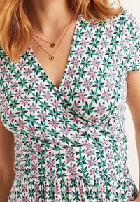 Bloemenprint wikkeljurk van witte stof, met een groen, roze en marineblauw patroon, een ingezette taille en korte mouwen. Twee gelaagde gouden kettingen.