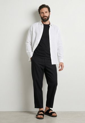 Mand med skæg iført hvid skjorte med knapper over en sort T-shirt, sorte bukser til knæene og sorte sandaler, står op ad en almindelig hvid væg.