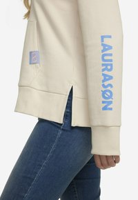 Cremefarvet sweatshirt med blå tekst, der står "LAURASØN," med en sideslids og en lomme, der er detaljeret med et pink og blåt emblem.
