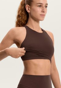 Barna, rugalmas sportmelltartó magas nyakkal, versenyző hátú dizájnnal és testhez simuló derékrésszel. Az anyag puha, sima textúrájú, minimális varrásokkal.