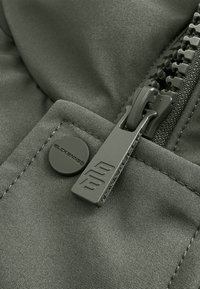 Close-up van donkere stof met een metalen ritssluiting en een ronde knop, beide voorzien van de logo's "BLACKBANN5" en "22".