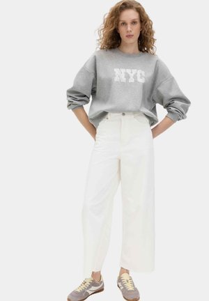 Junge Frau mit lockigem Haar, die einen grauen NYC-Sweatshirt, weiße weitgeschnittene Hose und beige Sneakers trägt und mit den Händen in der Hüfte steht.